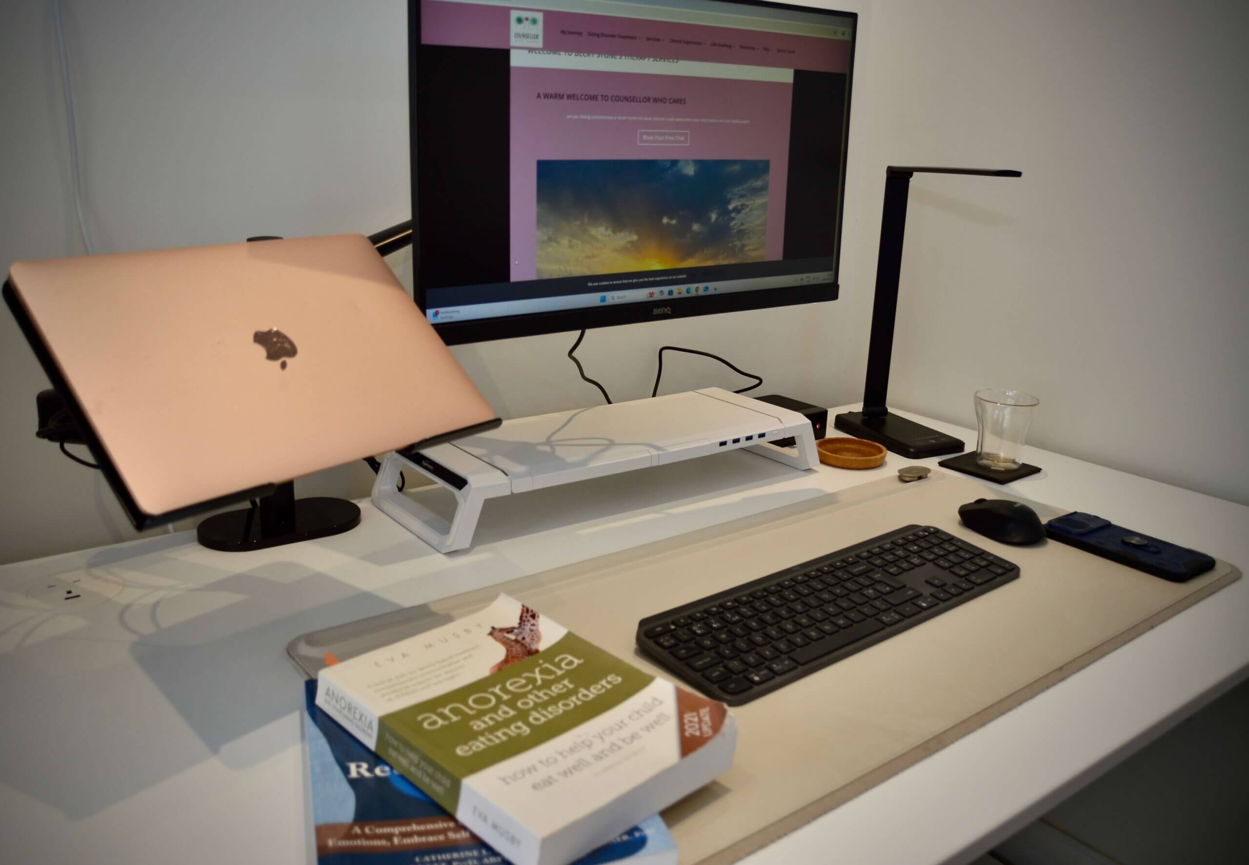 Becky Stone's home office desk set up for online counselling sessions, with a laptop, monitor showing the Counsellor Who Cares website, and eating disorder recovery books including Anorexia and Other Eating Disorders by Eva Musby
